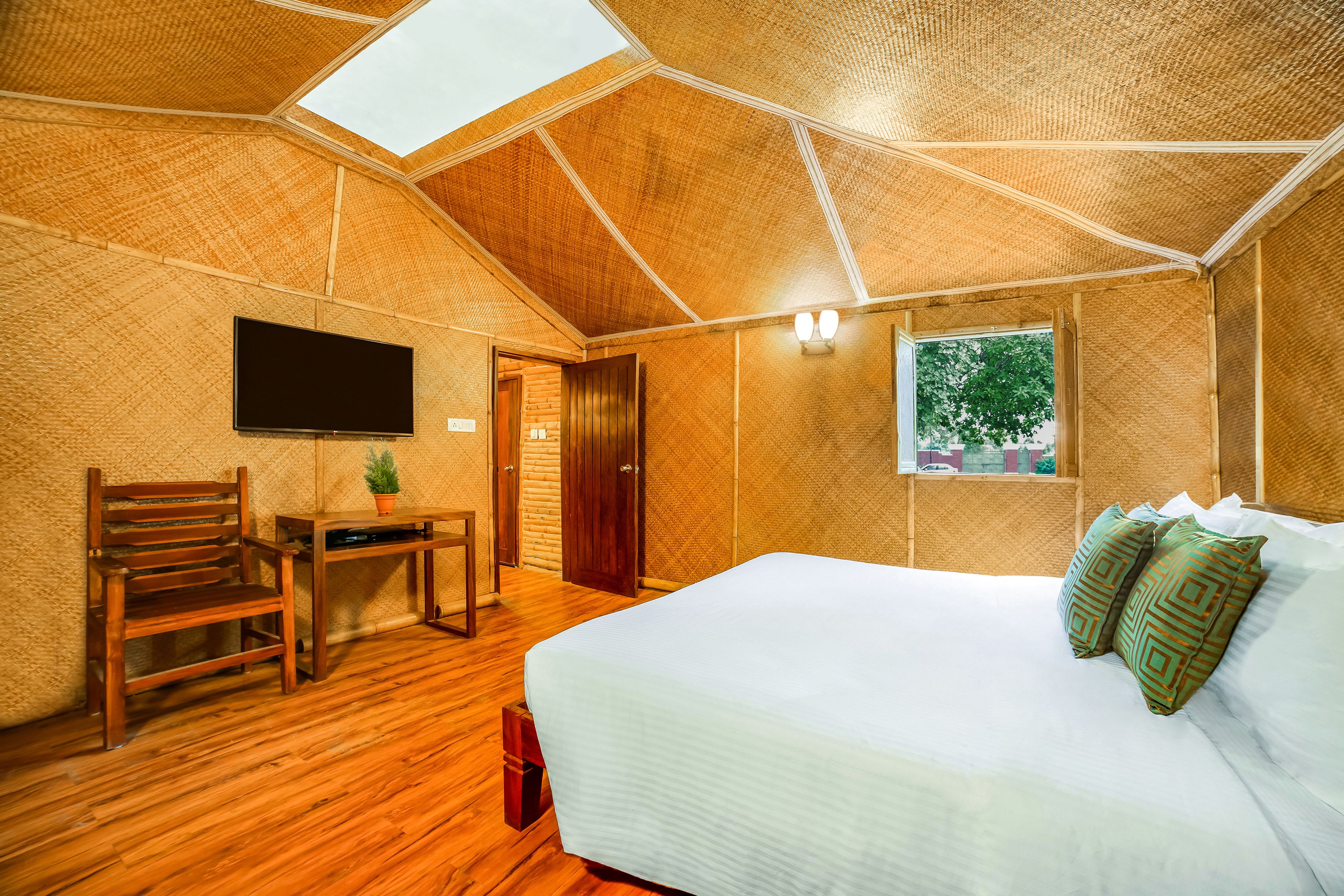 A room with a sloped ceiling, a bed with patterned pillows, a flat-screen TV, a wooden chair, a small table with a plant, and a window with a view of trees.