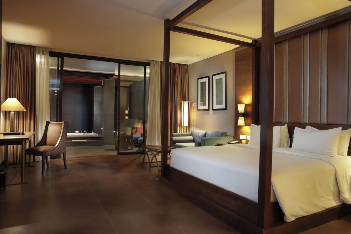 Habitación de hotel con una cama grande, una zona de estar con sofá y sillones, un escritorio con silla y una puerta de cristal que da a un balcón.