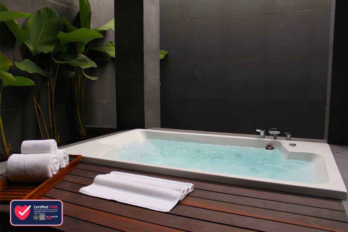 Una bañera de hidromasaje rectangular con un deck de madera en primer plano, un par de toallas blancas y una planta verde grande al fondo.