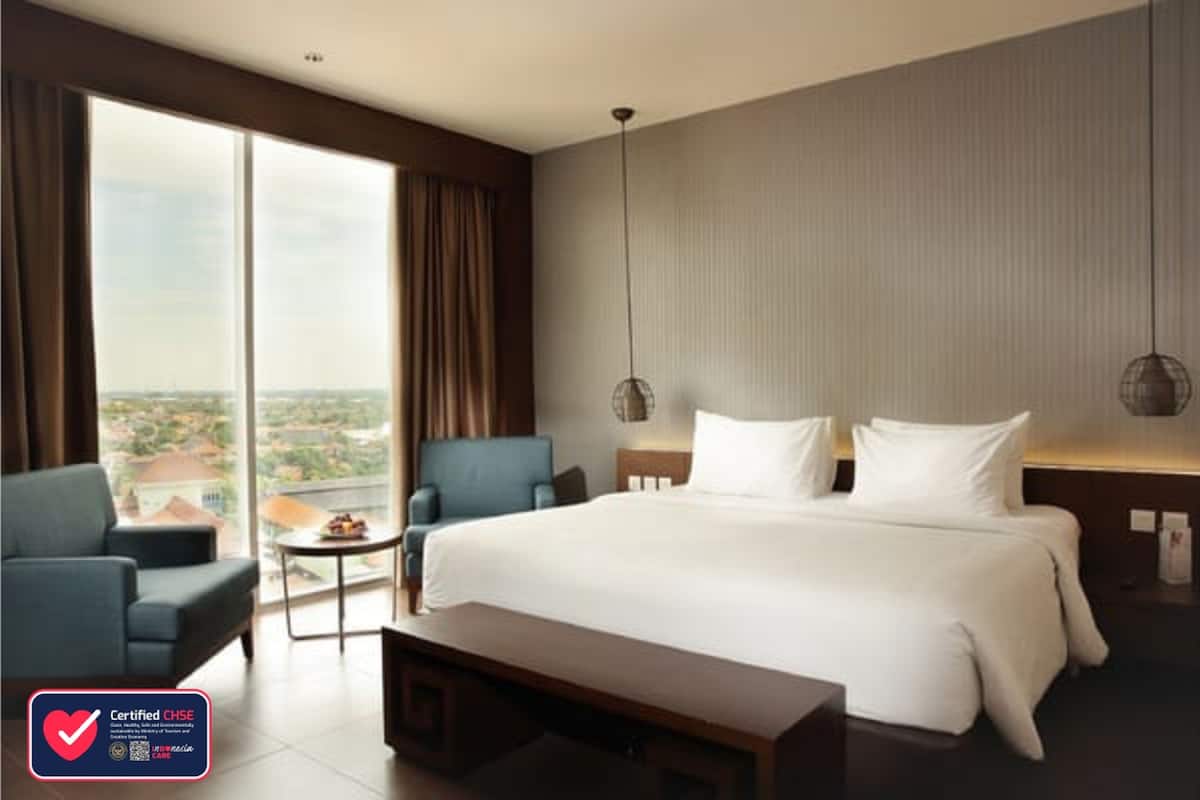 Habitación de hotel con una cama grande, un sofá, una mesita y vistas a la ciudad a través de amplios ventanales.