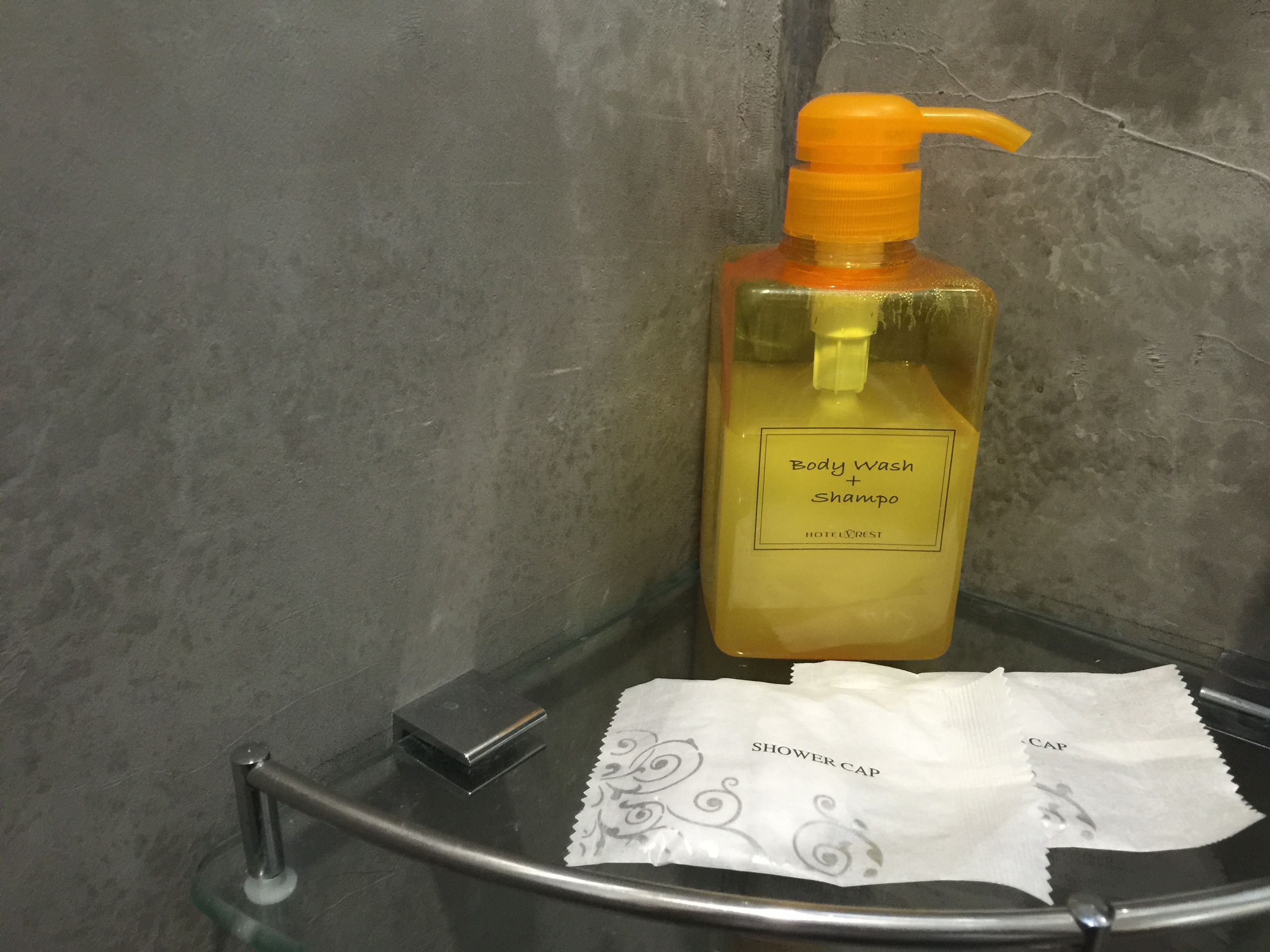 A bottle of body wash and shampoo, a shower cap, and a small black object on a glass shelf in a bathroom.