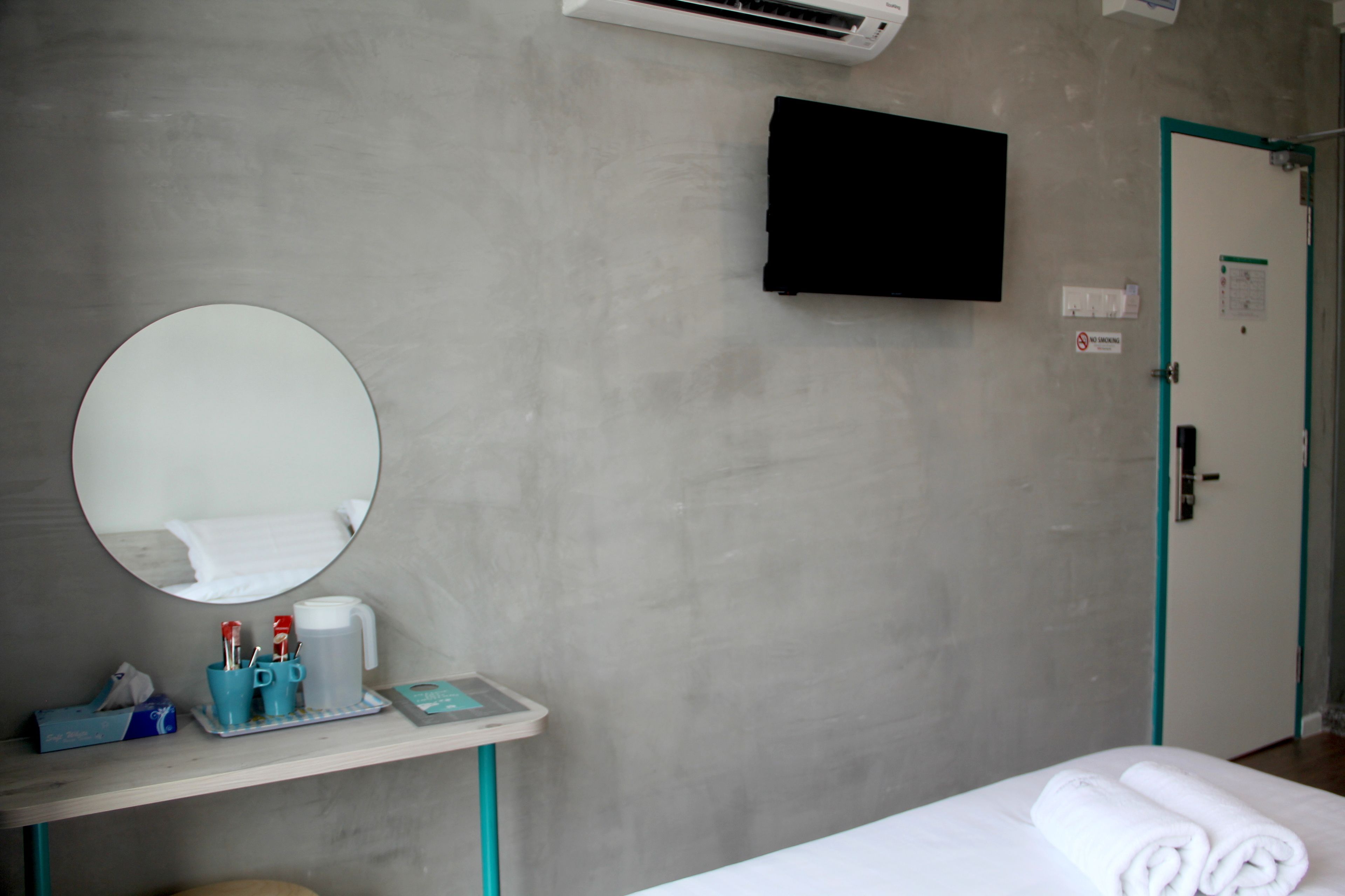 A hotel room with a bed, a round mirror, a small table with toiletries, a TV mounted on the wall, and a door with a green frame.