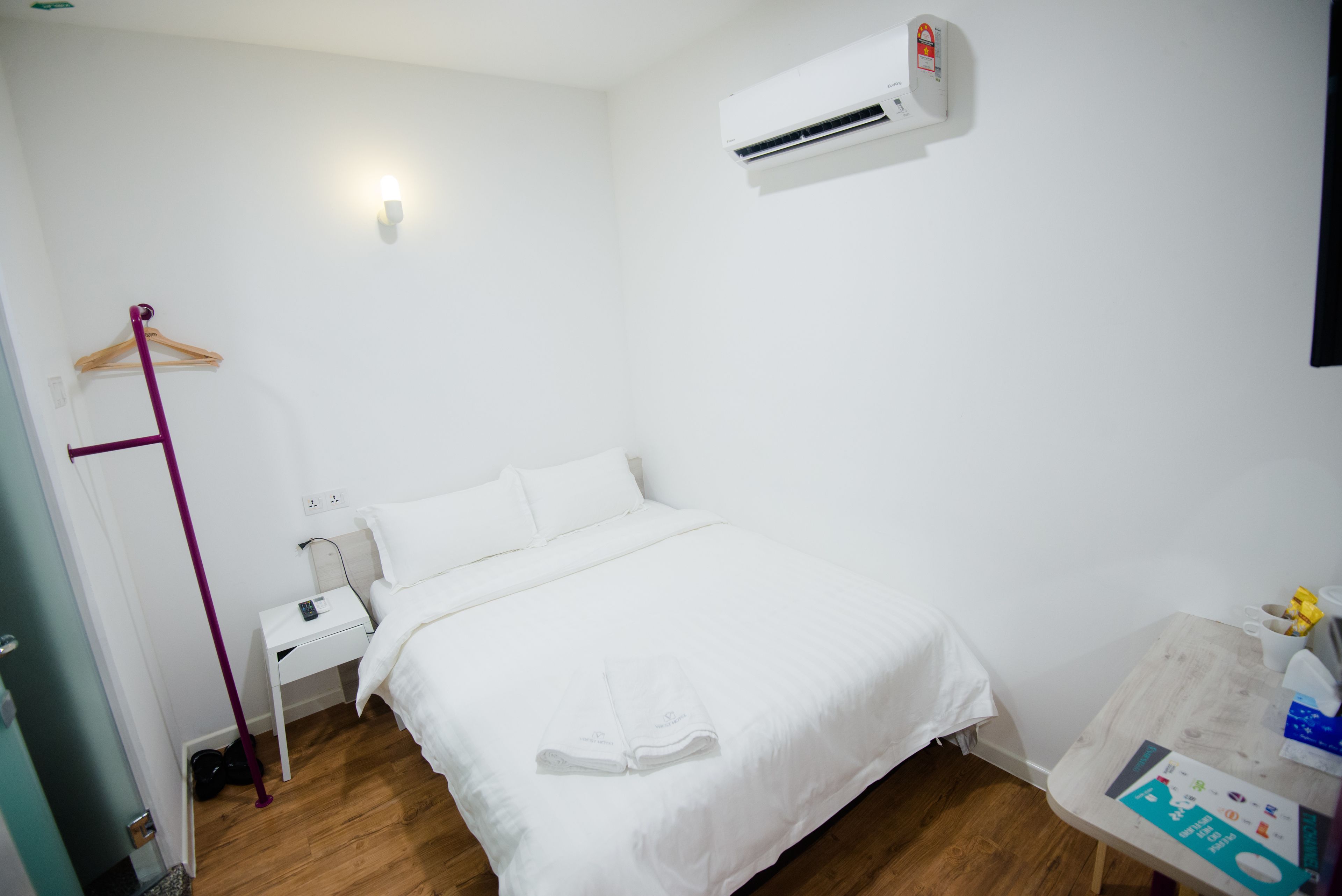A clean, white hotel room with a bed, bedside table, and a wall-mounted air conditioner.