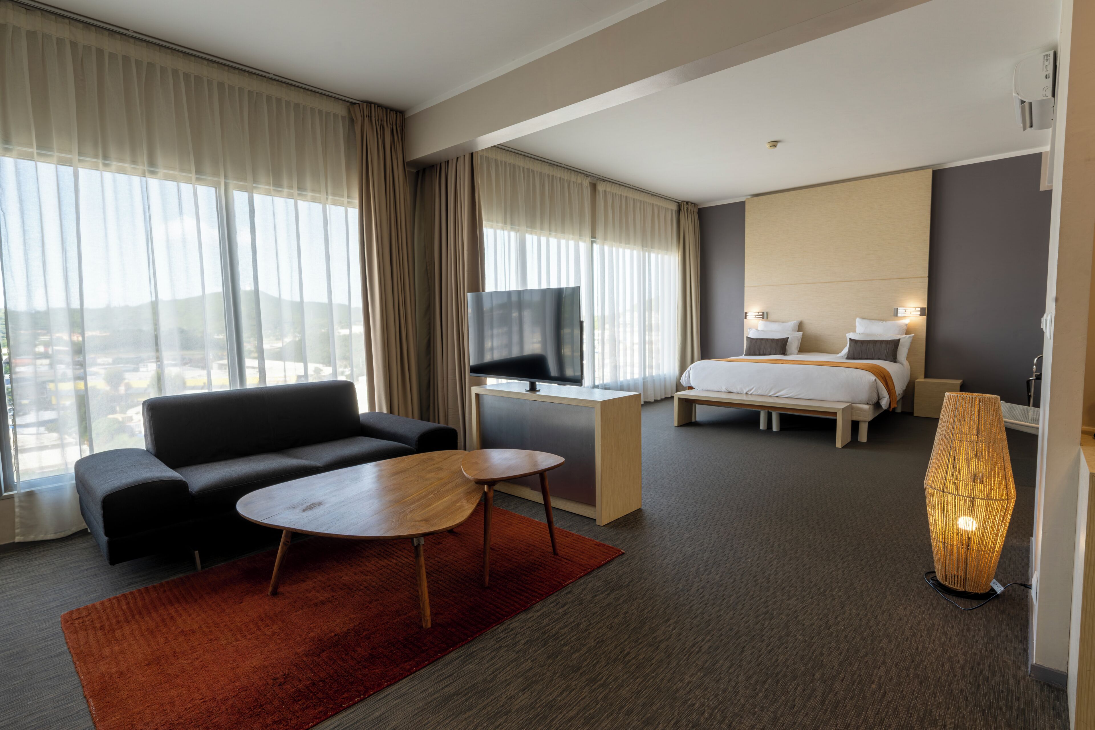 A hotel room with a sofa, a small table, a bed, a desk with a TV, and a large window with curtains.
