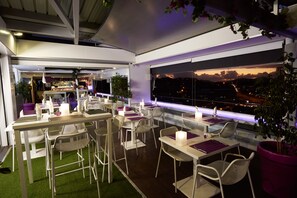 Un área de comedor al aire libre con mesas y sillas, un bar al fondo y vistas de la ciudad al anochecer.