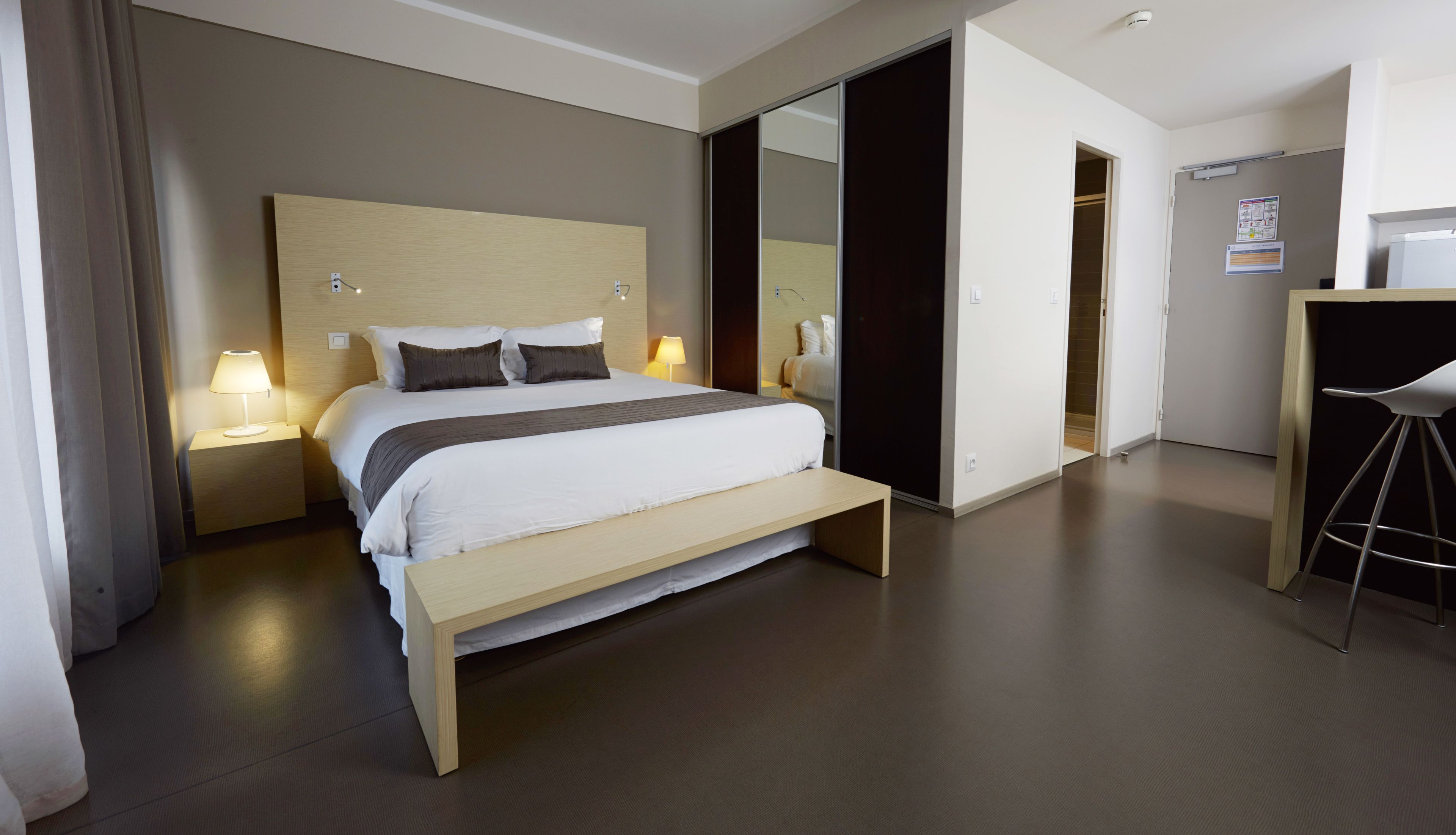 A modern hotel room with a large bed, bedside lamps, a wooden bench, a desk with a chair, and a mirror.
