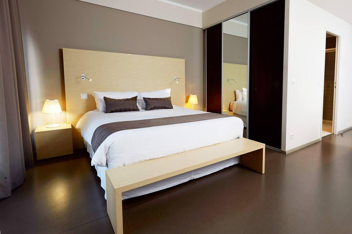 A modern hotel room with a large bed, bedside lamps, a wooden bench, and a sliding door leading to a bathroom.