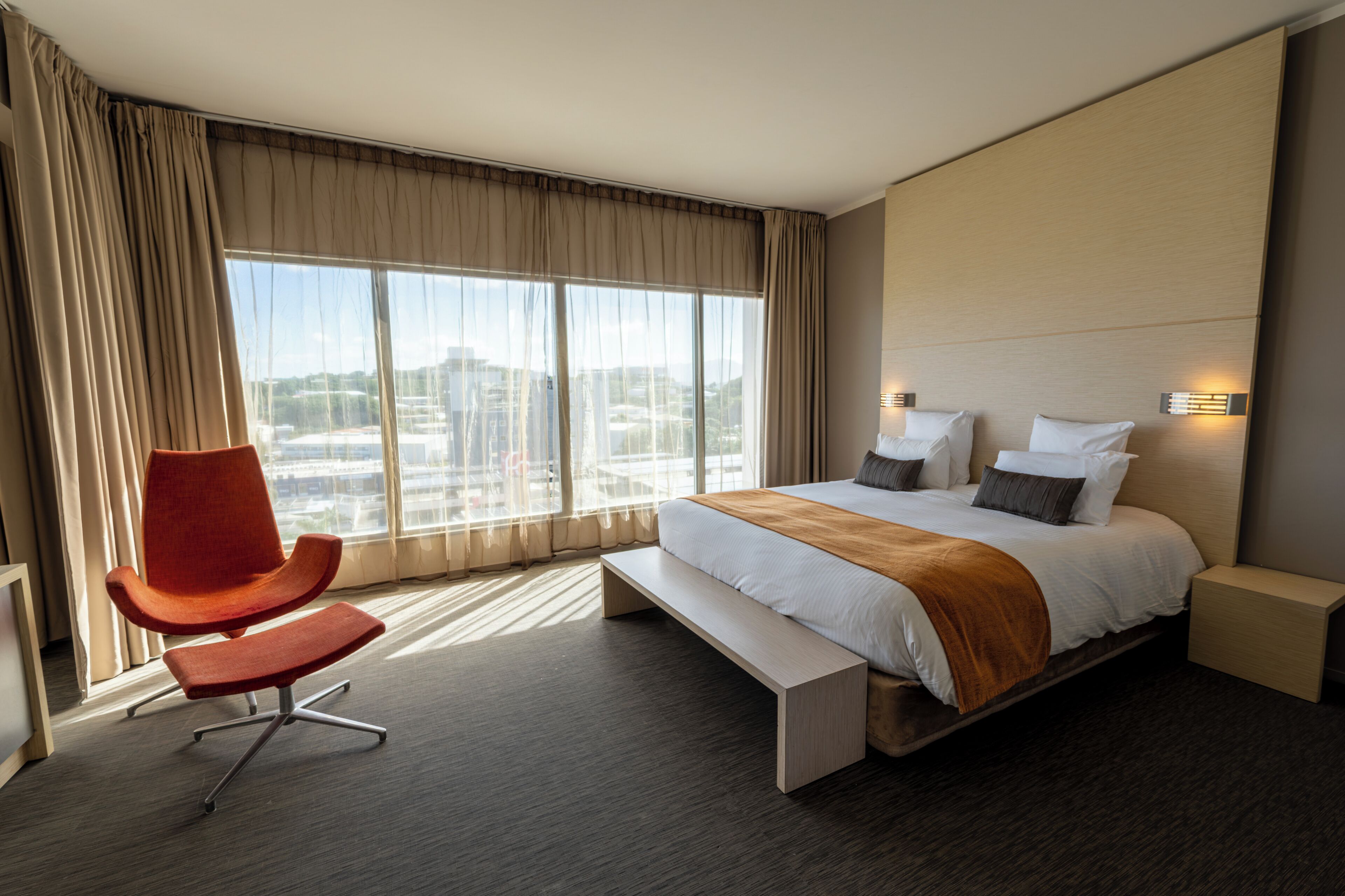 A hotel room with a large bed, a bench, a red chair, and a view of the city through the window.