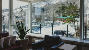 A room with a large window offering a view of a snowy outdoor area, a cushioned seating area, and a potted plant. - Mountainside Hakuba (Hakuba)