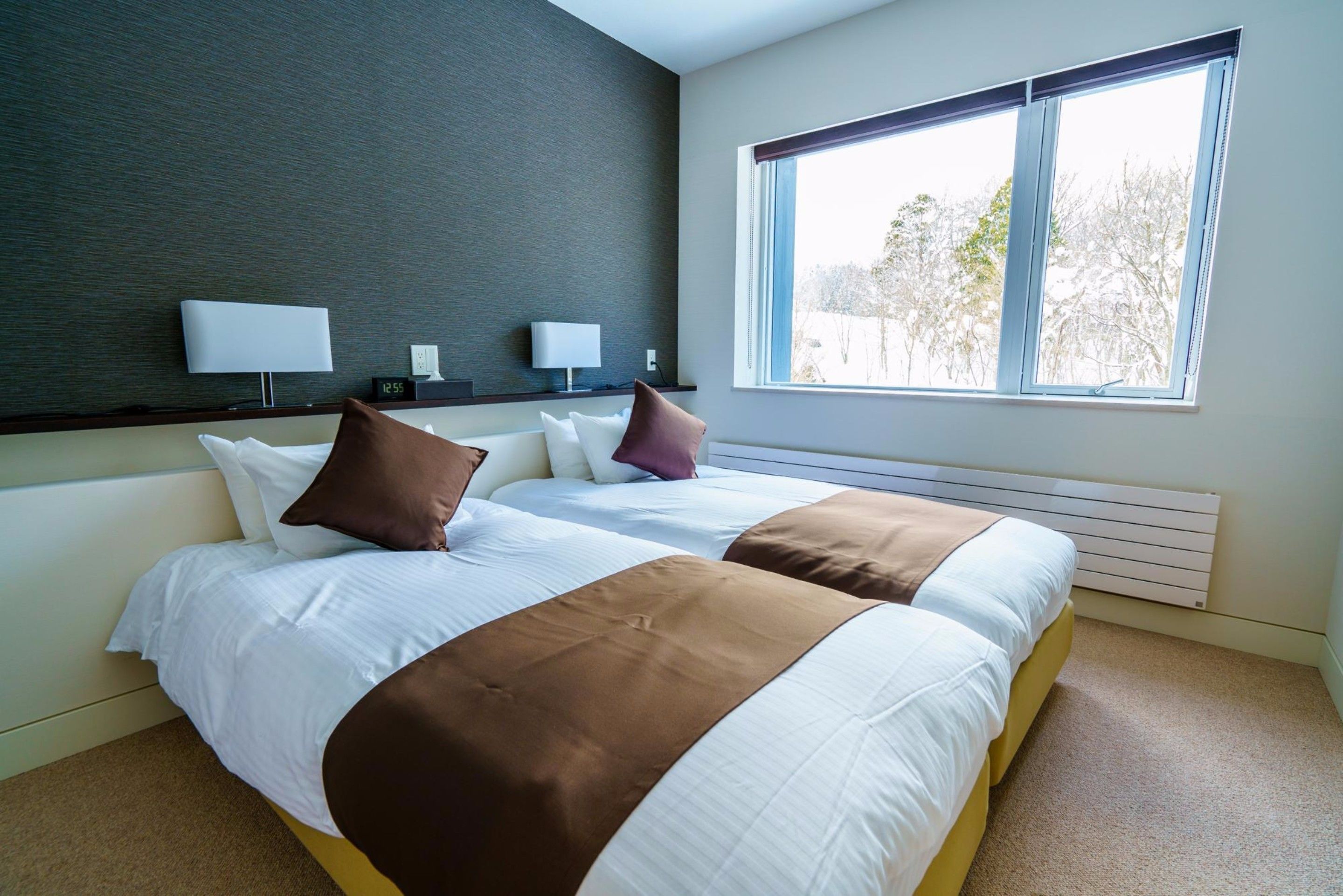 A hotel room with a large bed, two bedside tables with lamps, a window with a view of trees, and a dark wall.