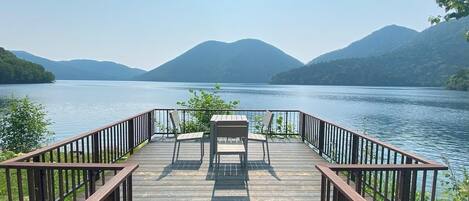A wooden deck with a table and chairs overlooking a lake with mountains in the background.