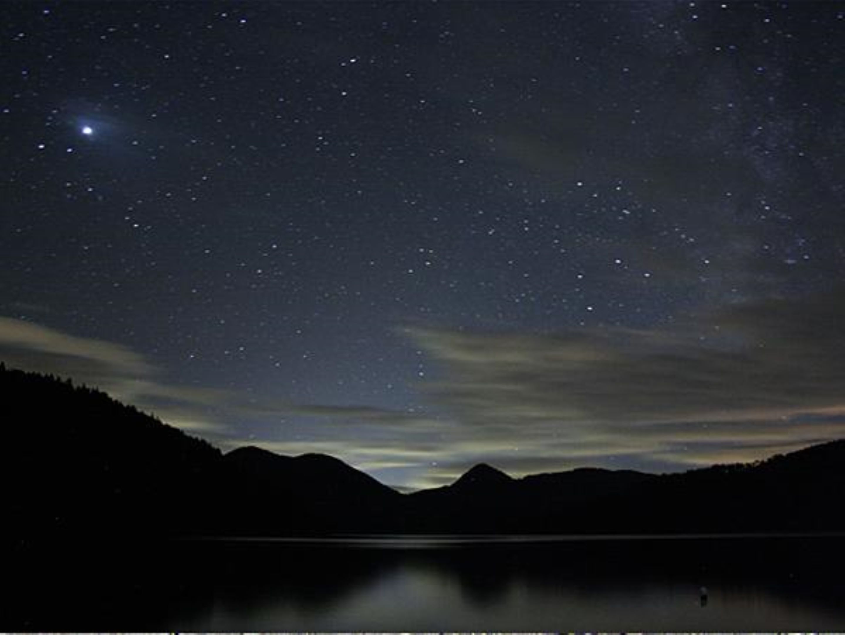 A night sky filled with stars over a calm lake with mountains in the background.