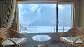 A room with two wicker chairs and a glass table, a large window with a view of mountains and a body of water. - Shikaribetsu Lakeside Onsen Hotel Fusui (Shikaoi)