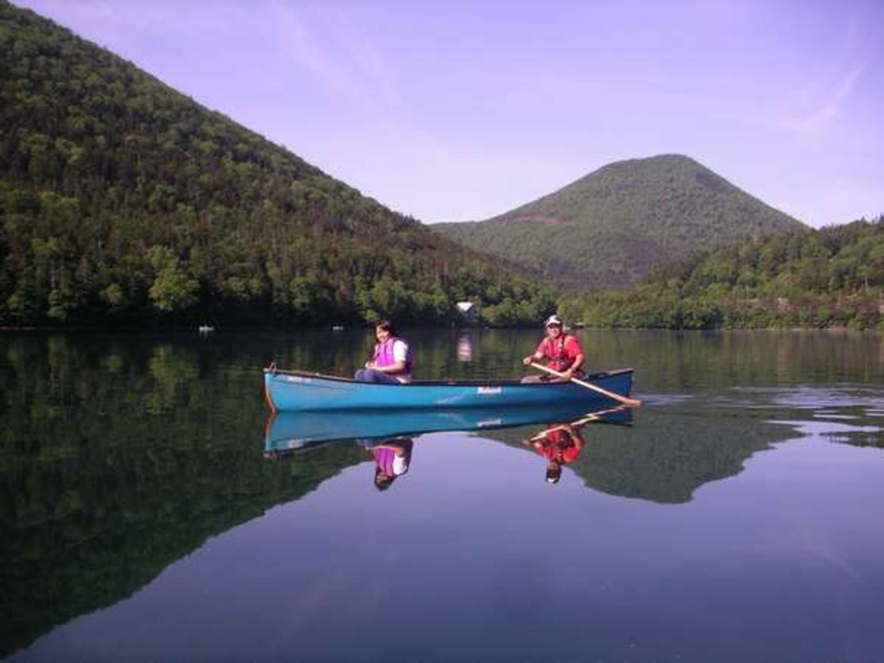 A calm lake with two people in a blue canoe, surrounded by forested hills.