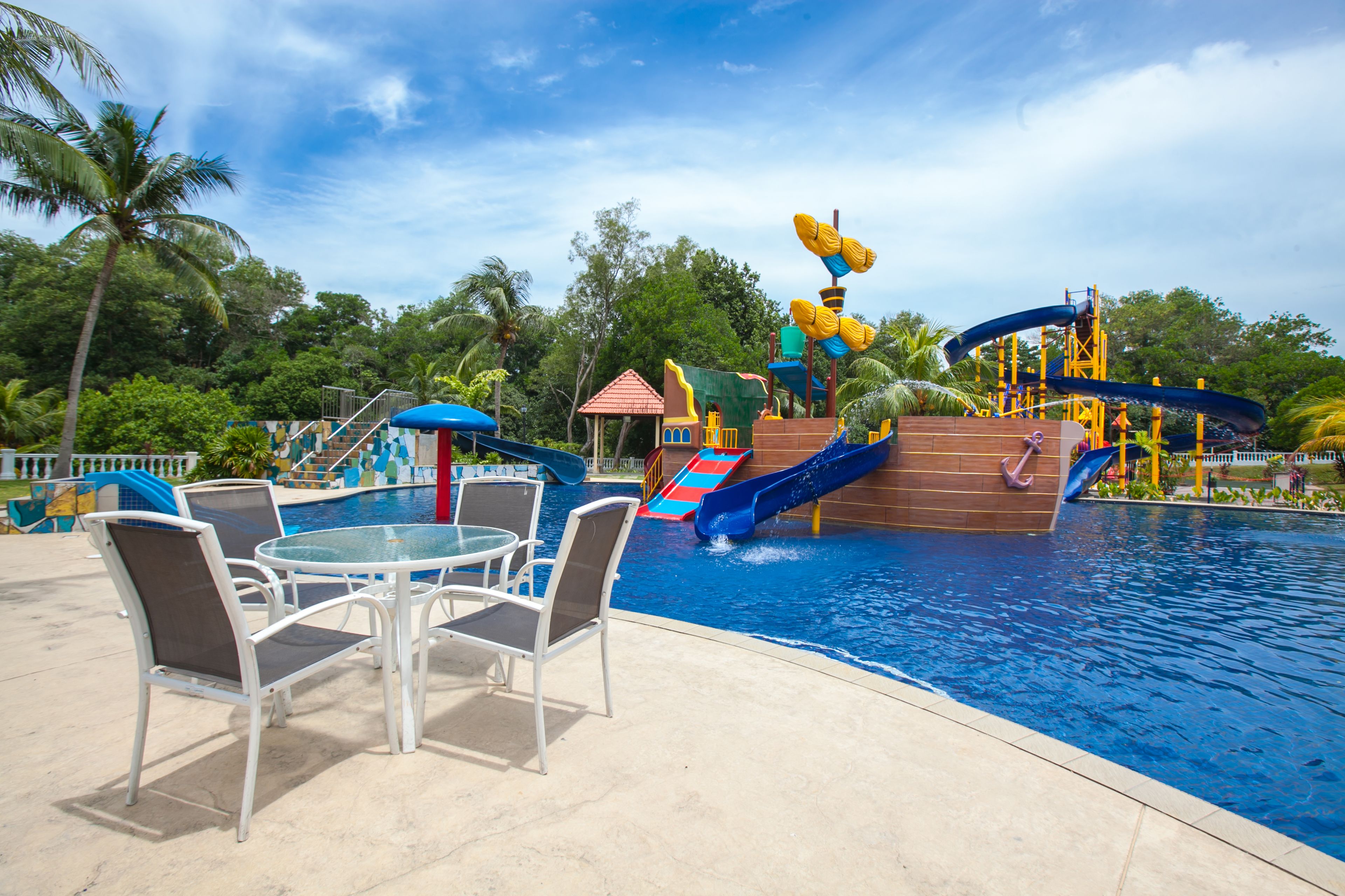 A pool area with chairs and a table, a water slide, and a small building.