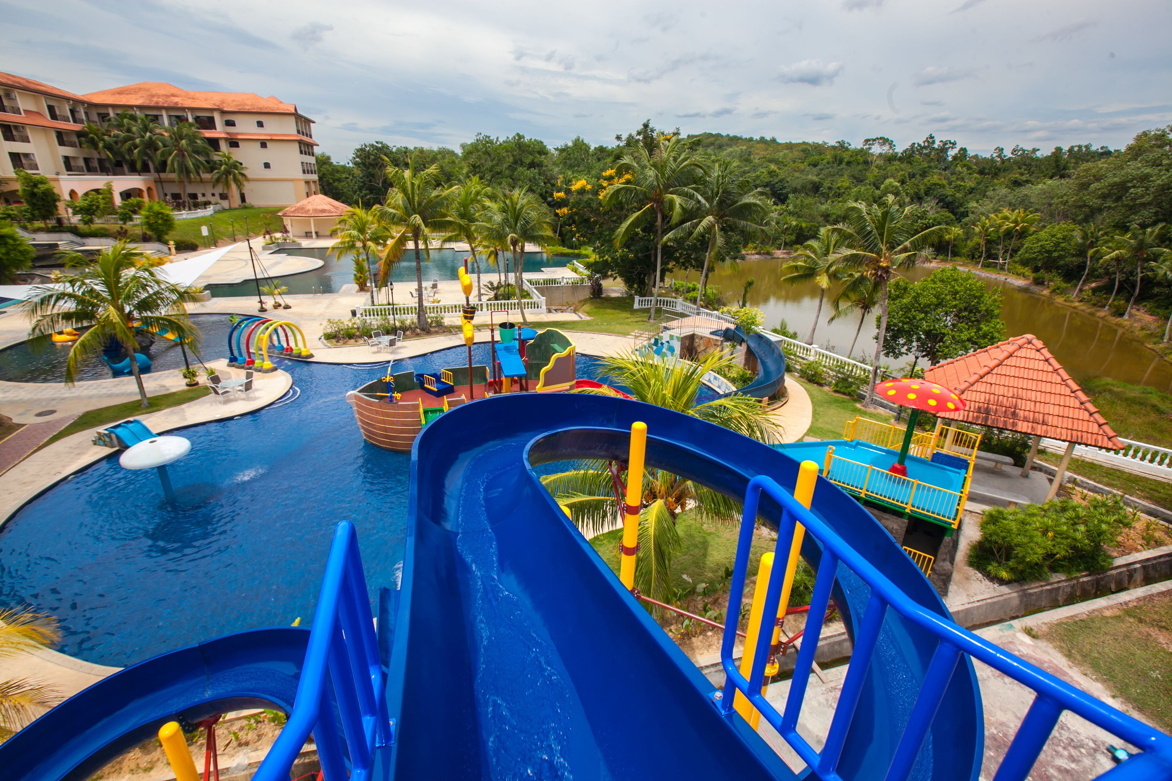 A water park with multiple slides and pools, surrounded by palm trees and a large building in the background.