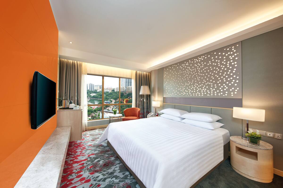 Quarto de hotel moderno com uma cama grande, uma TV de tela plana, uma escrivaninha com luminária e vista para a cidade através de amplas janelas.
