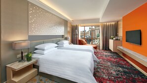 A hotel room with two beds, a TV, a desk, and a view of the city.