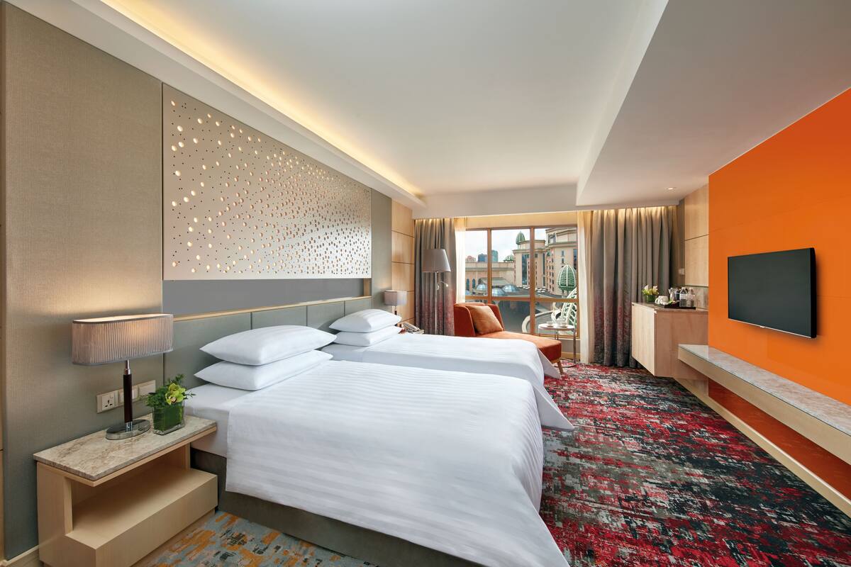 Quarto de hotel com duas camas, televisão, escrivaninha e vista para a cidade.
