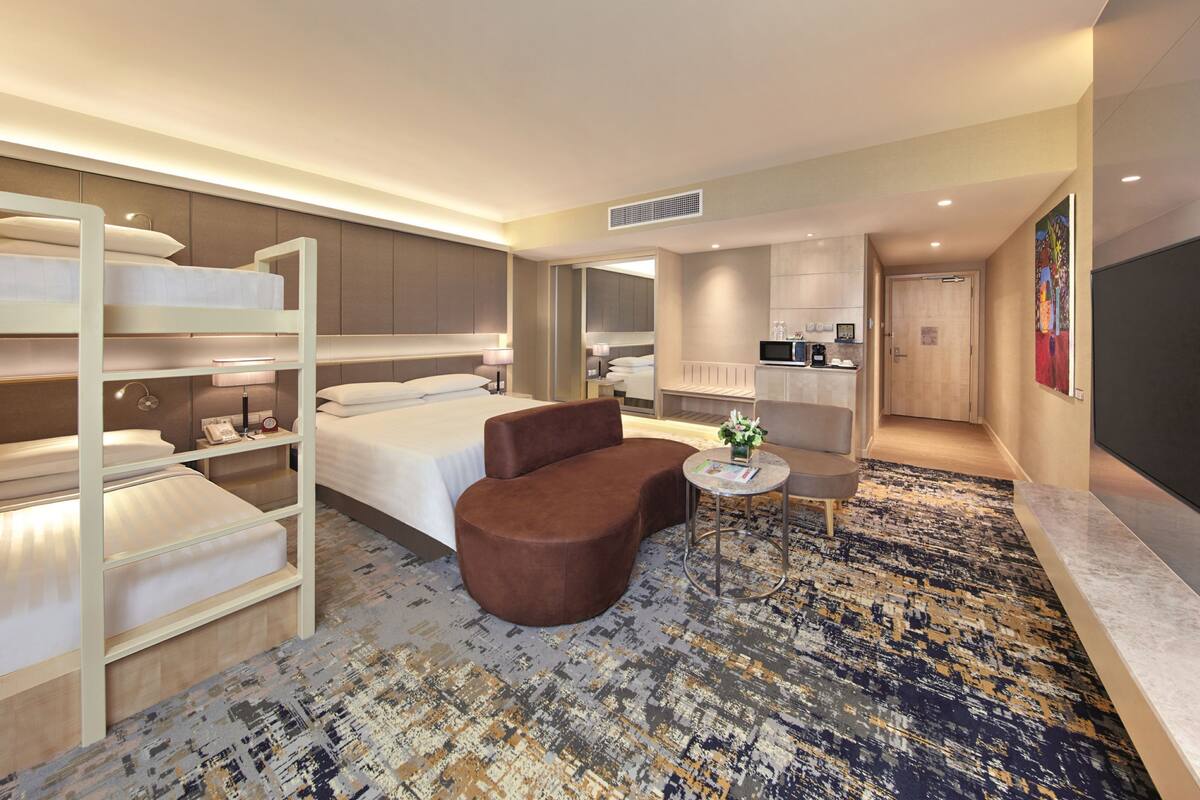 Quarto de hotel com cama, sofá, mesinha e cozinha compacta.