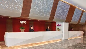 A modern hotel reception area with a white reception desk, two receptionists, and decorative potted plants.
