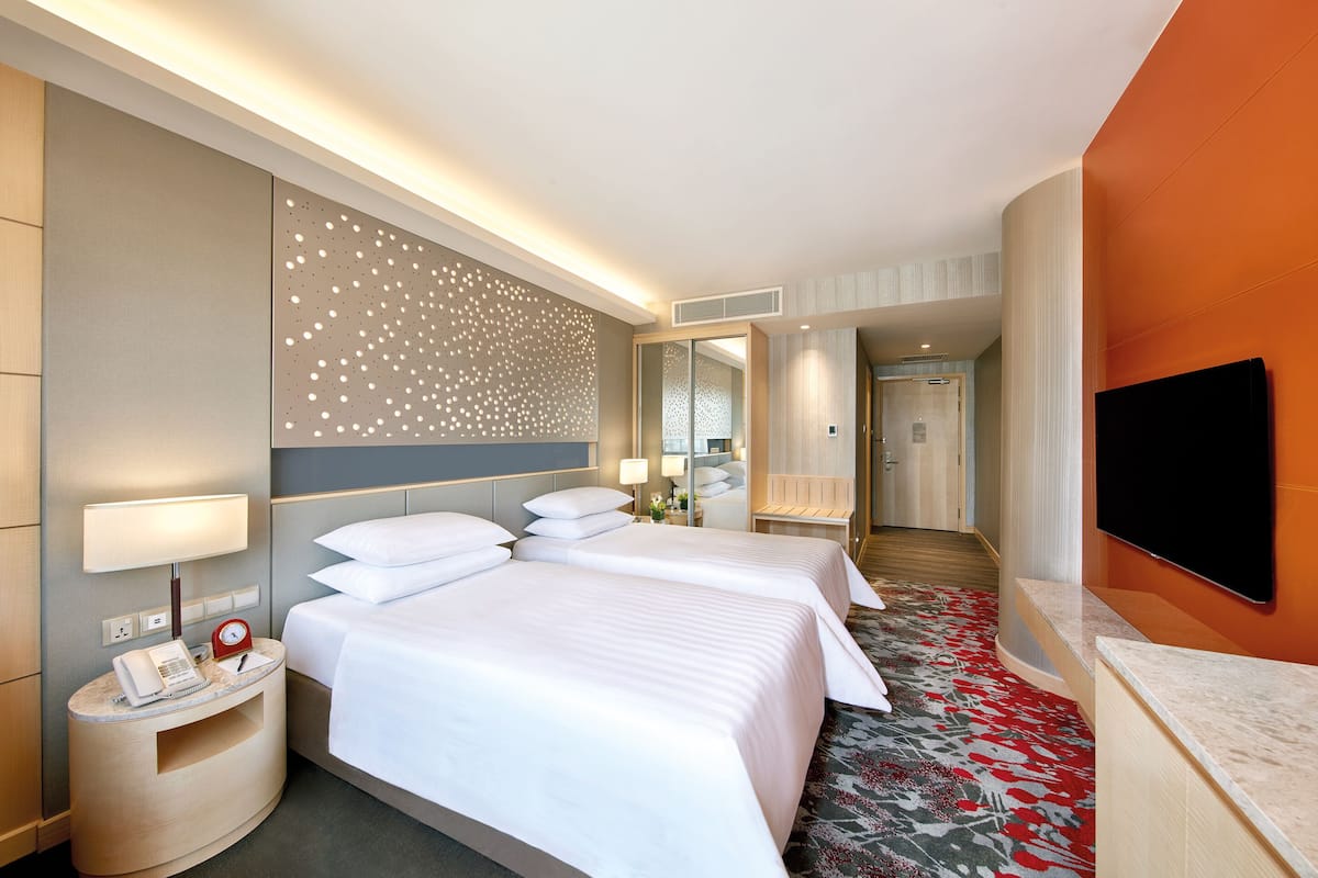 Quarto de hotel com duas camas, uma escrivaninha com abajur, telefone e televisão na parede.