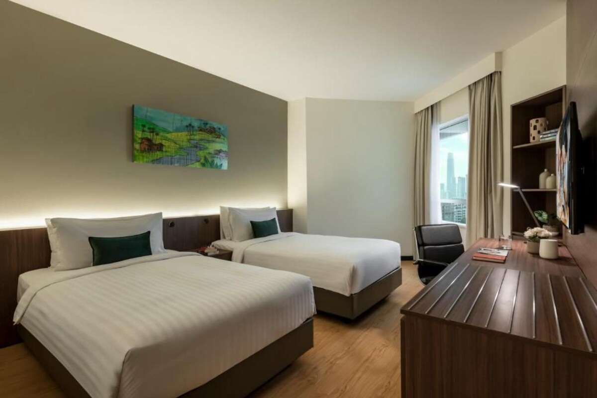 A hotel room with two beds, a desk, a chair, a painting on the wall, and a window with curtains.