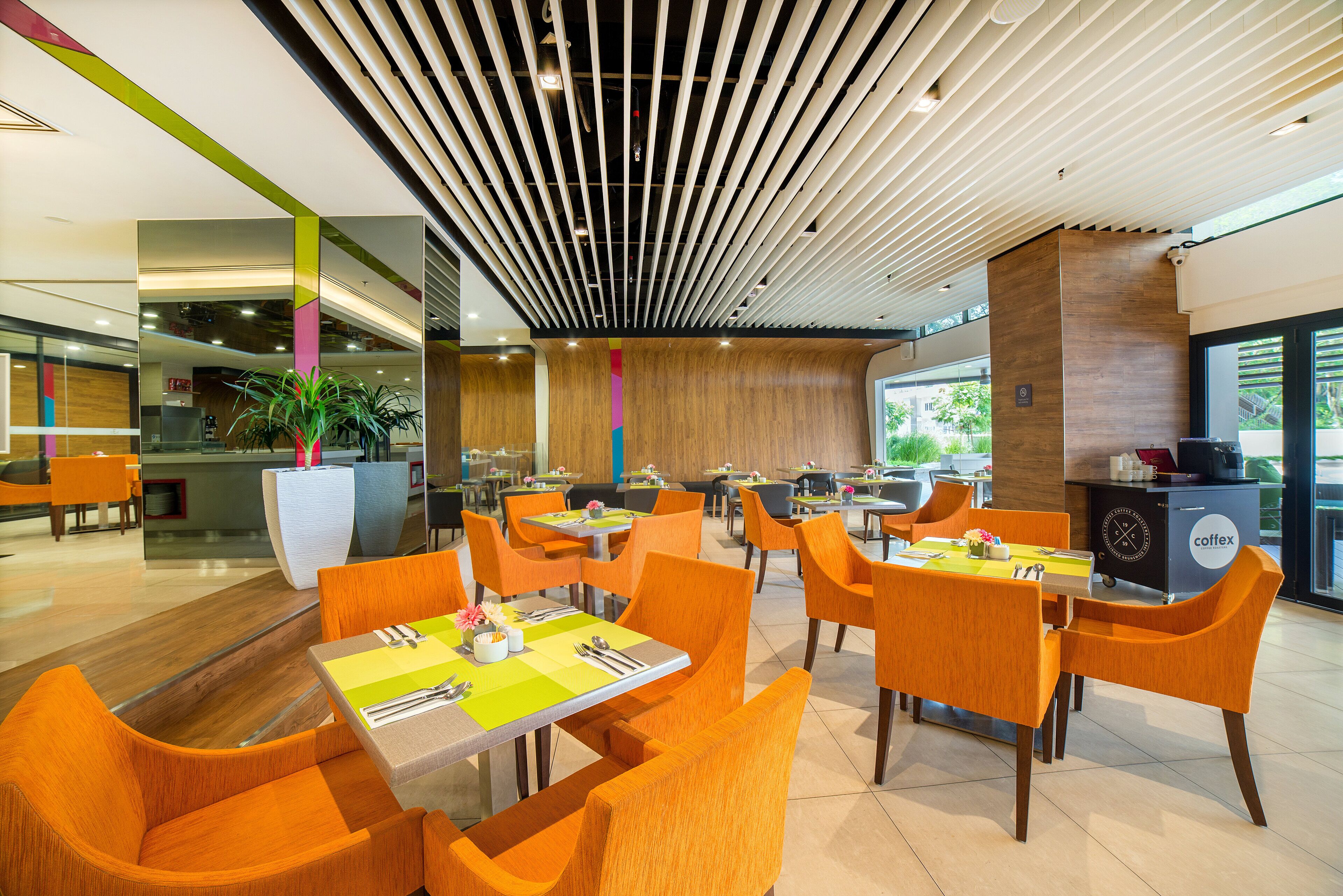 Un restaurant moderne avec des chaises et des tables orange, un mur d’accent en bois et un plafond doté d’un éclairage linéaire.
