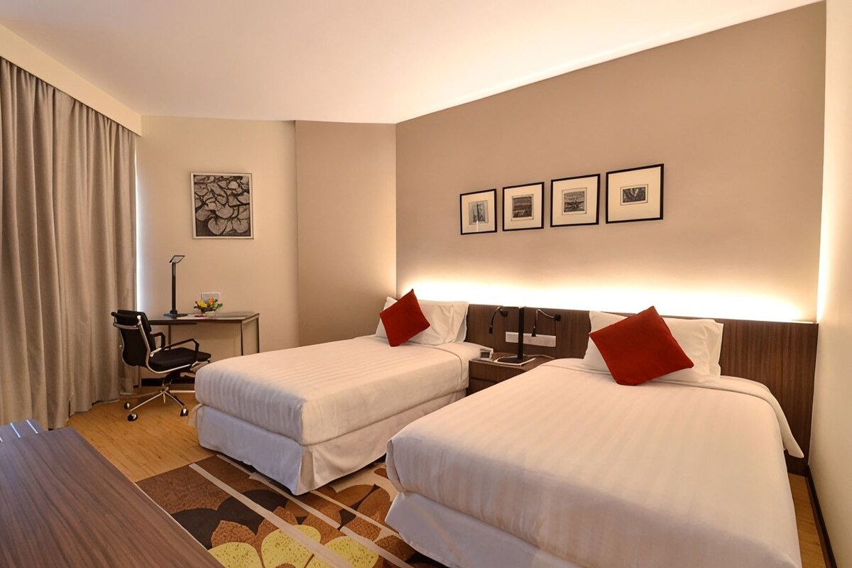 A hotel room with two beds, a desk, a chair, and a lamp.
