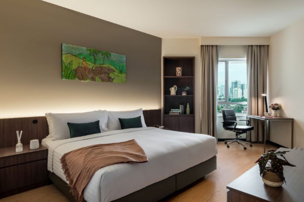 A hotel room with a large bed, a desk, a chair, a bookshelf, and a painting on the wall.