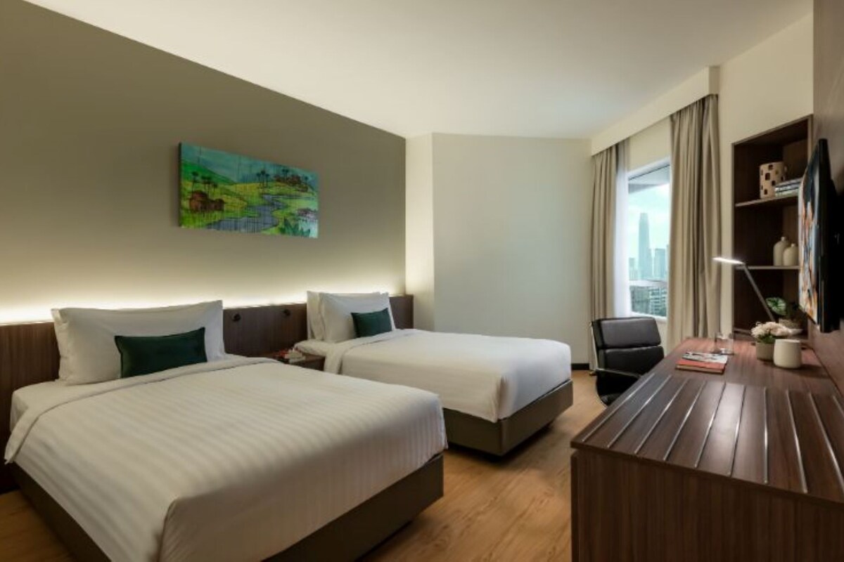A hotel room with two beds, a desk, a chair, a painting on the wall, and a window with a city view.