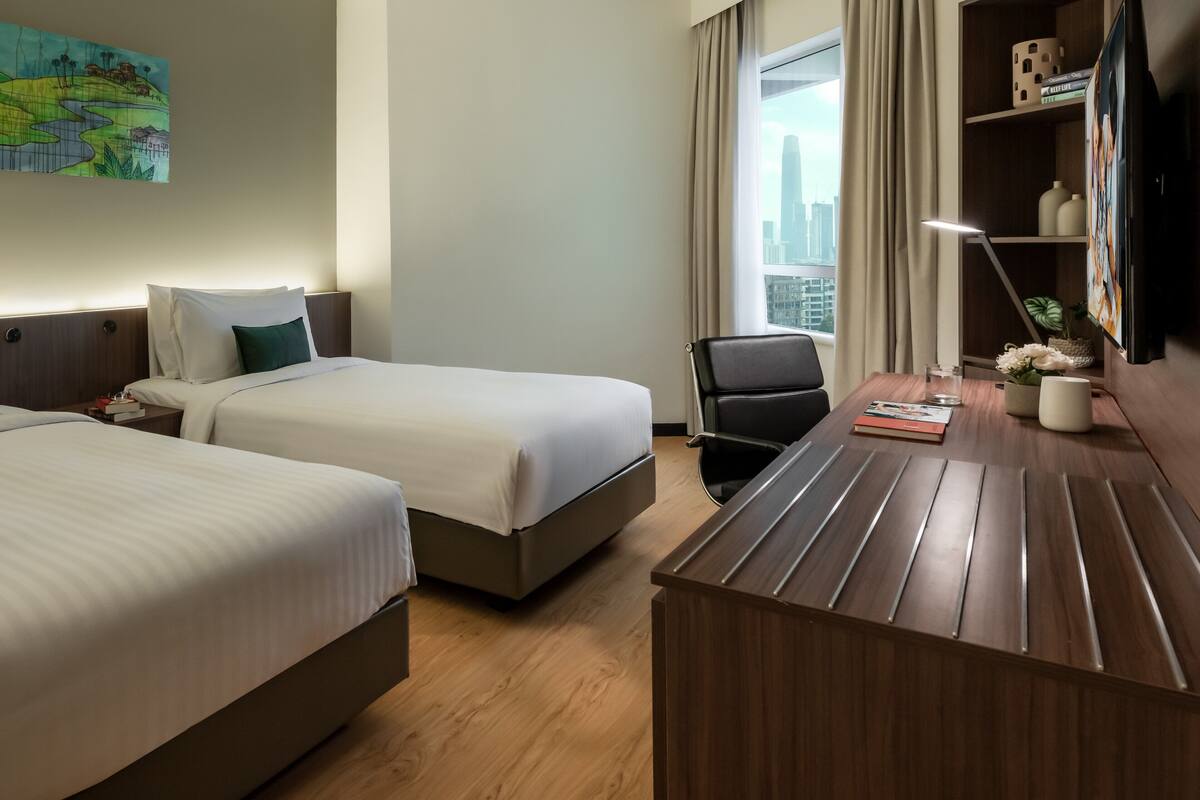 A hotel room with two beds, a desk, a chair, a lamp, a bookshelf, and a window with a city view.
