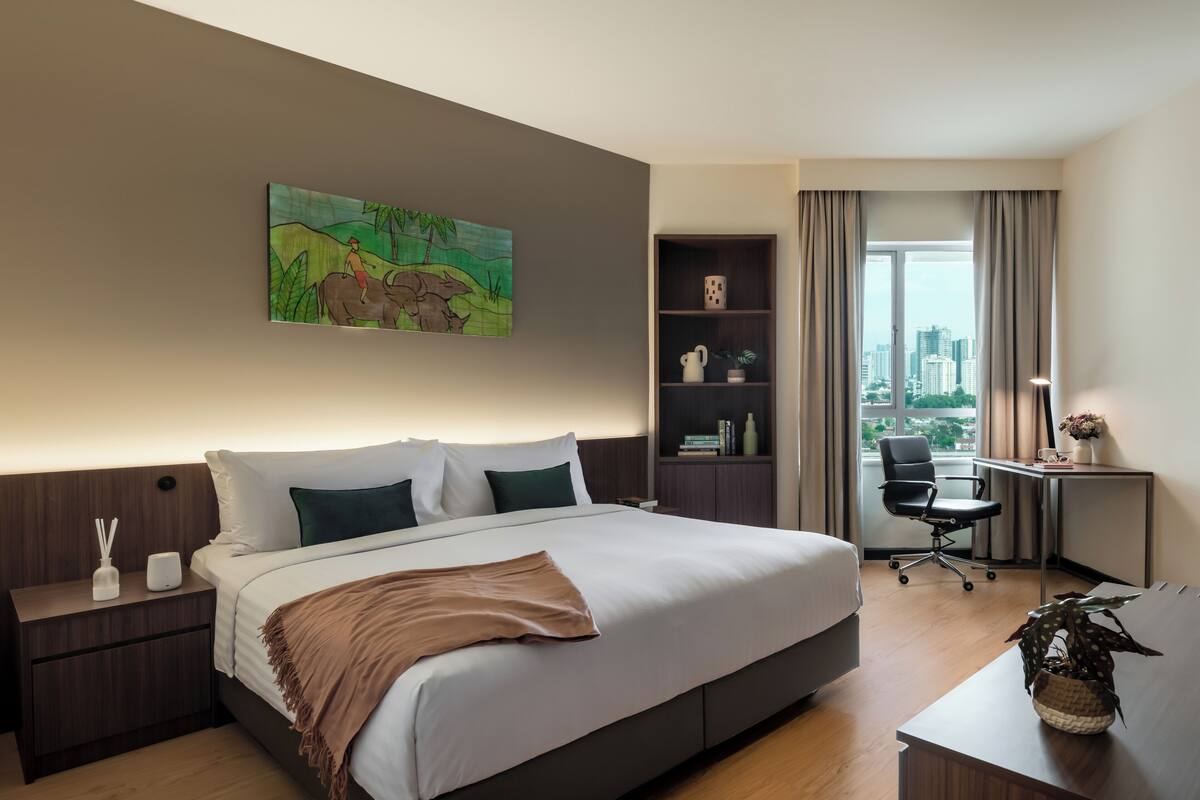 A hotel room with a large bed, a desk, a chair, a bookshelf, and a painting on the wall.