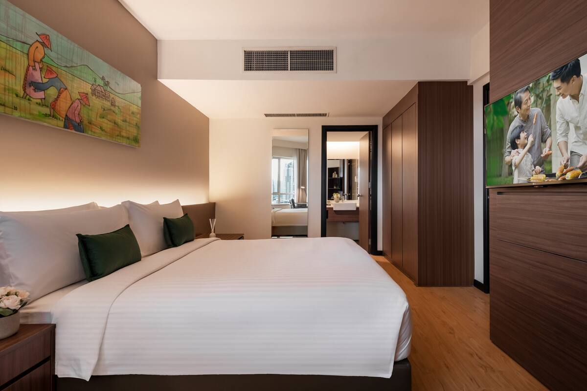 A hotel room with a large bed, a painting on the wall, a television, and a view into the bathroom.