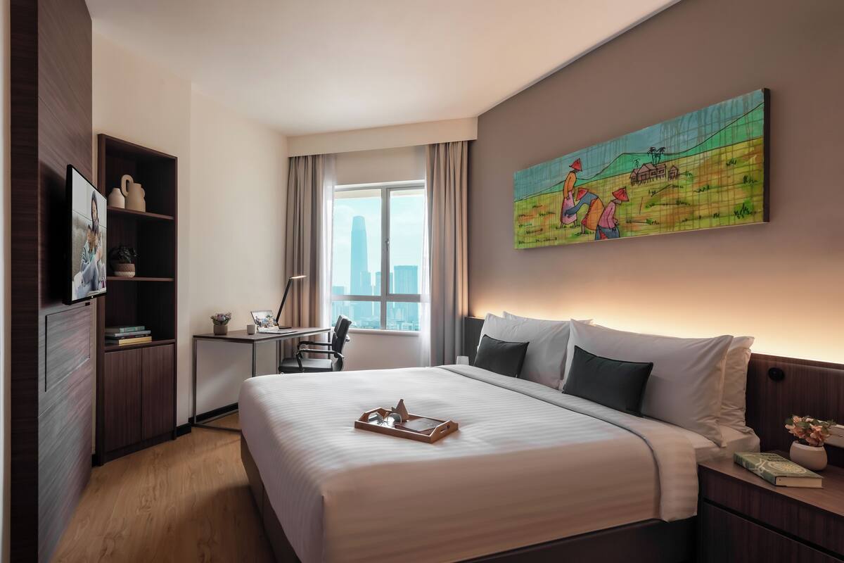 A hotel room with a large bed, a desk, a chair, a bookshelf, and a painting on the wall.