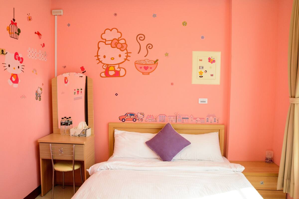 Quarto de hotel em tons de rosa, com cama, escrivaninha de madeira, cadeira e cômoda.