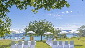 An outdoor wedding setup with chairs and umbrellas, a large tree, and a lake view.