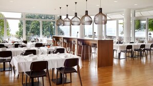 A spacious restaurant with multiple tables set for dining, large windows offering a view, and modern pendant lighting.