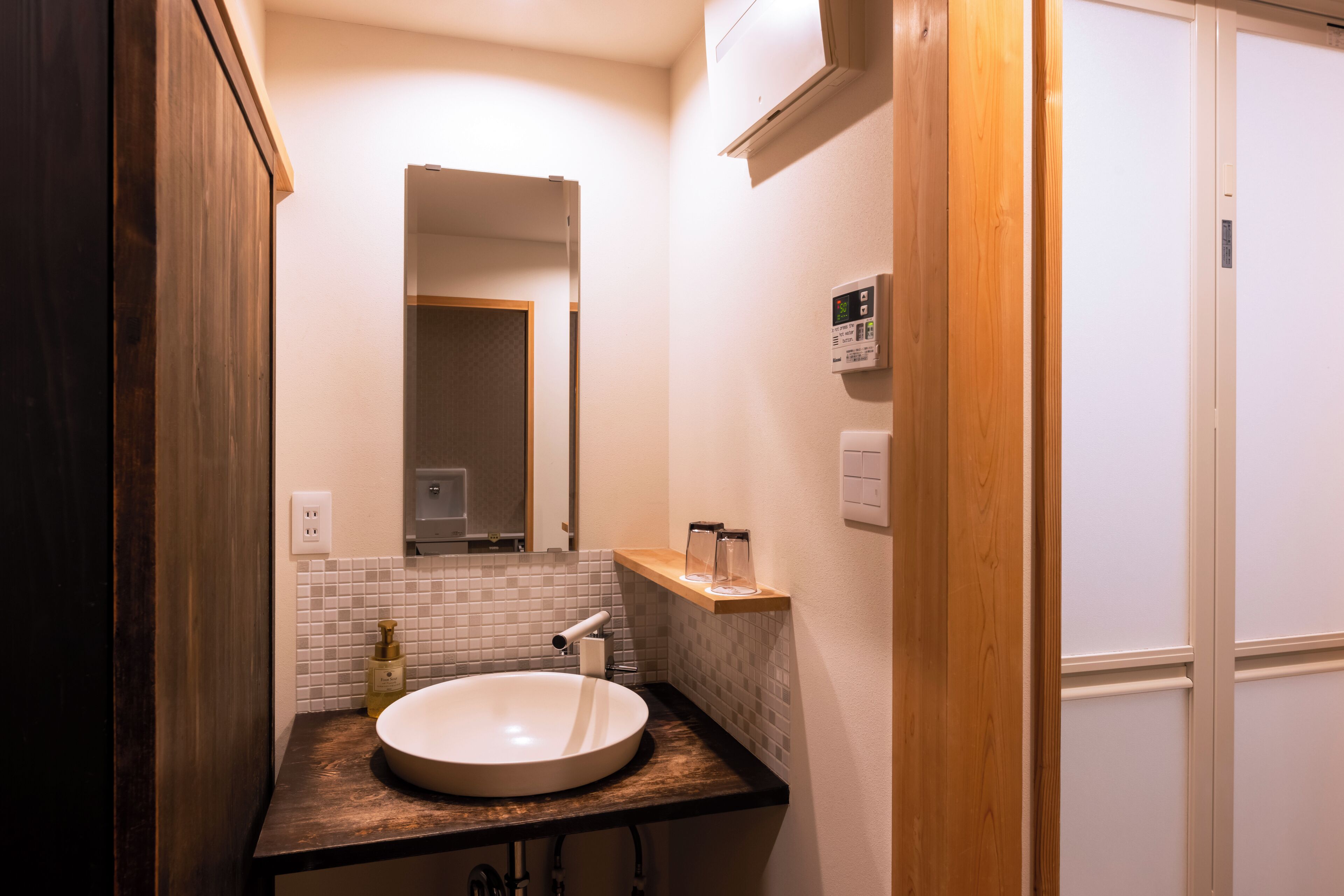 A bathroom with a round sink, a mirror, a towel dispenser, and a small shelf with toiletries.