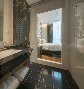 A modern bathroom with a glass shower enclosure, a large mirror, and a vanity with a sink and towel rack. - Luminor Hotel Pecenongan Jakarta by WH (Jakarta)