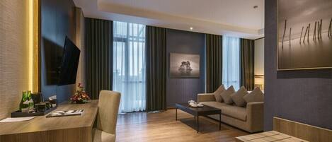 A modern hotel room with a wooden desk, a sofa, a coffee table, and a TV mounted on the wall.