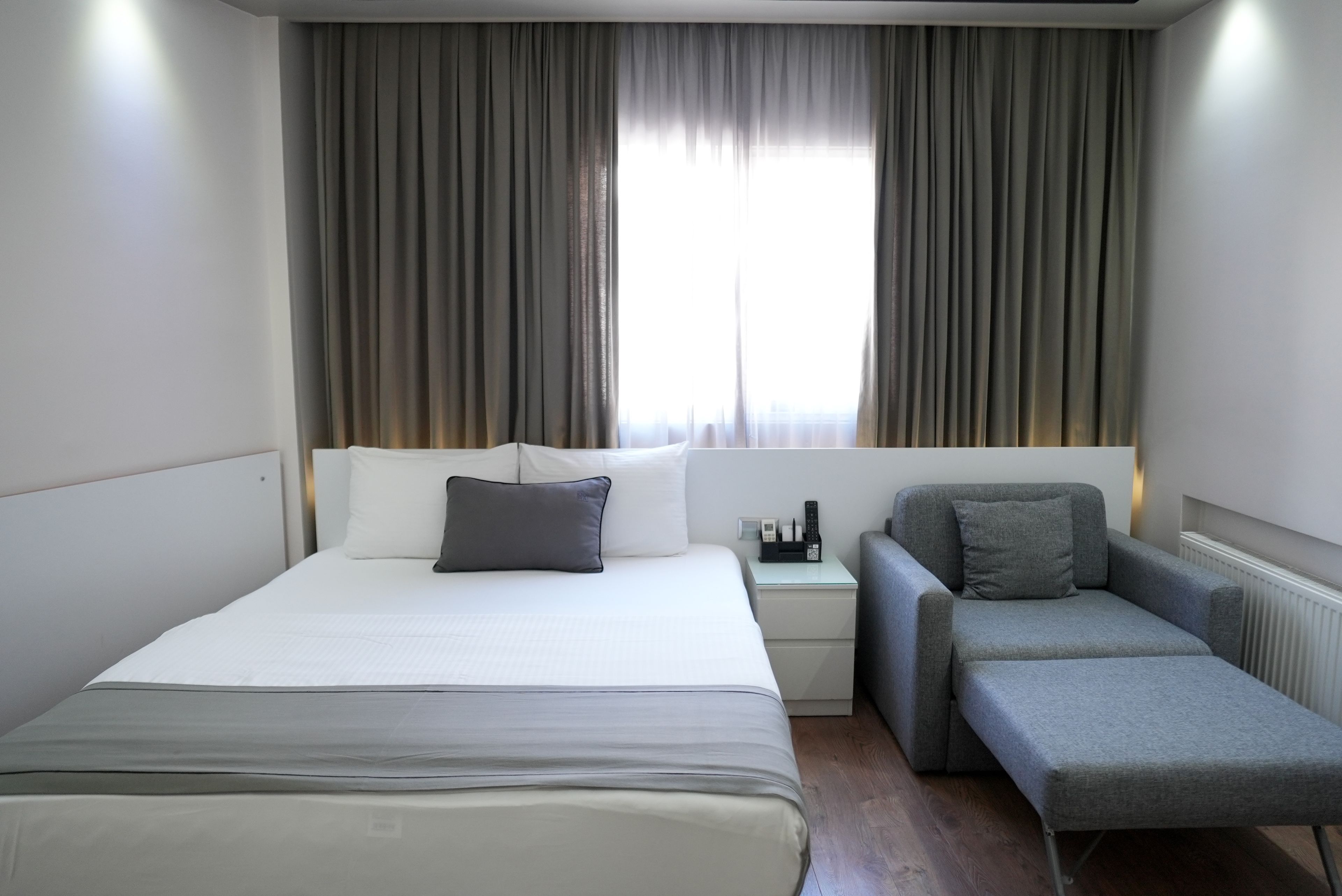 A hotel room with a bed, a chair, a bedside table with a lamp, and a window with curtains.