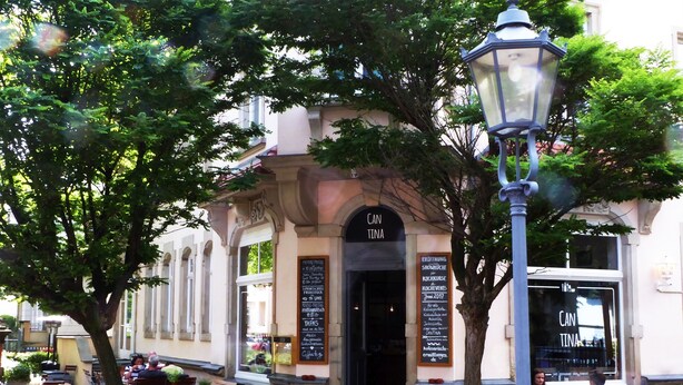 Ein Straßencafé mit Tischen und Stühlen, einer Straßenlaterne und einem Gebäudeeingang mit einem Schild.