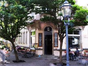 An outdoor café with tables and chairs, a street lamp, and a building entrance with a sign. - Ferienwohnung Weissbach (Dresden)