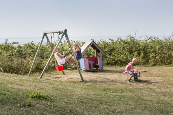 A swing set and a small playhouse in a grassy area with a view of the sea.