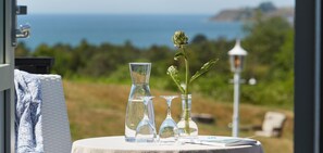 A table with clear glass vases and a single potted plant, set against a scenic view of the sea and greenery.