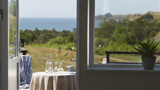 A room with a table set for dining, a potted plant on the windowsill, and a view of the ocean and greenery outside. - Löderups Strandbad Hotell & Stugby (Loderup)