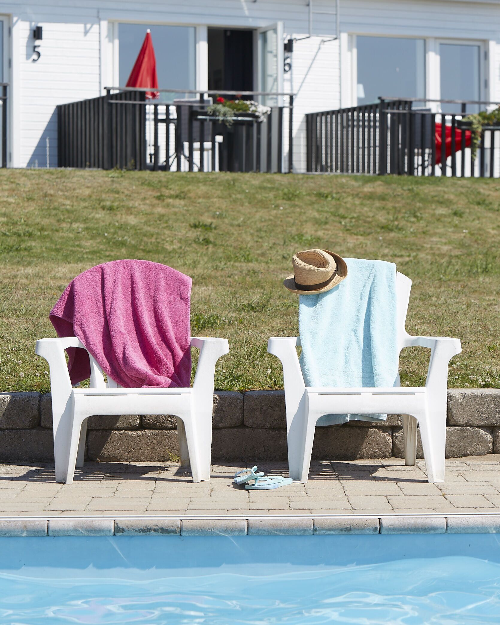 Two white plastic chairs by a pool with towels and a hat on them.