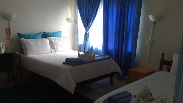 A hotel room with two beds, each with white bedding and blue accents. There are two bedside tables, one on each bed, and a small wooden nightstand with a lamp. The room has a tiled floor and a window with blue curtains. - Liya Guest Lodge (Kasane)