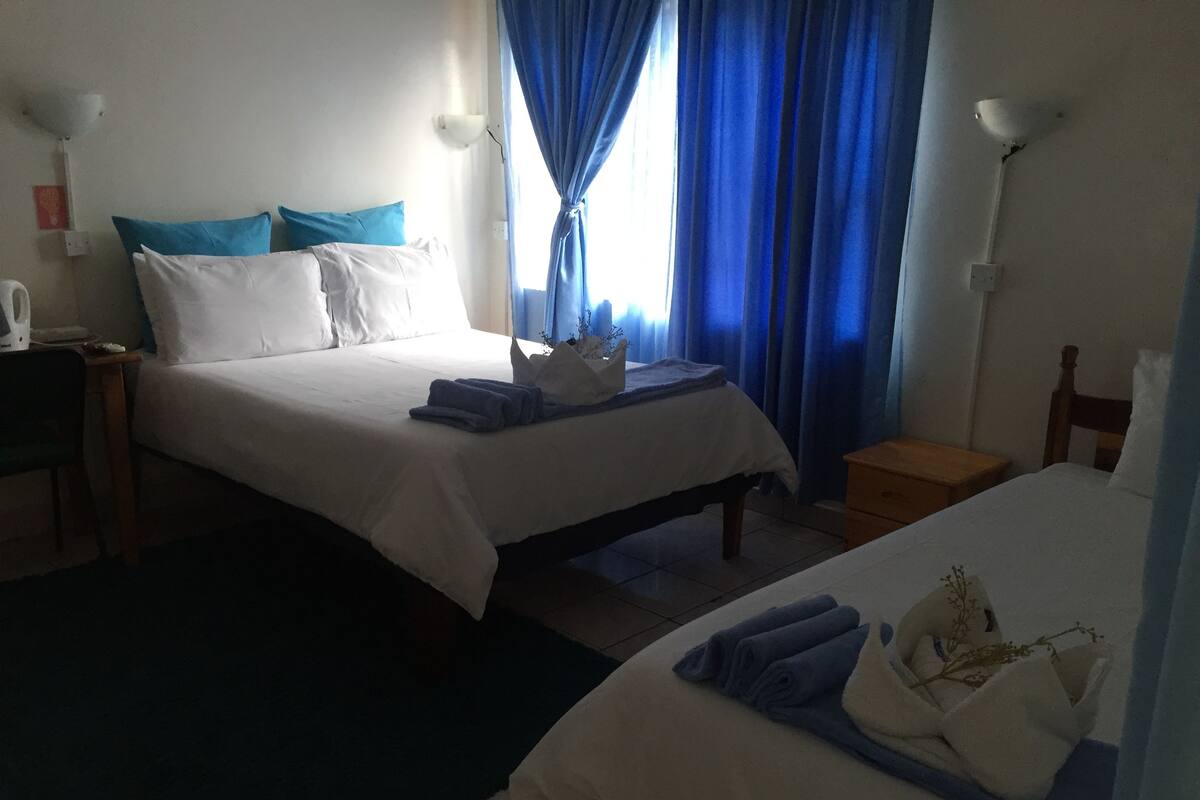 A hotel room with two beds, each with white bedding and blue accents. There are two bedside tables, one on each bed, and a small wooden nightstand with a lamp. The room has a tiled floor and a window with blue curtains.